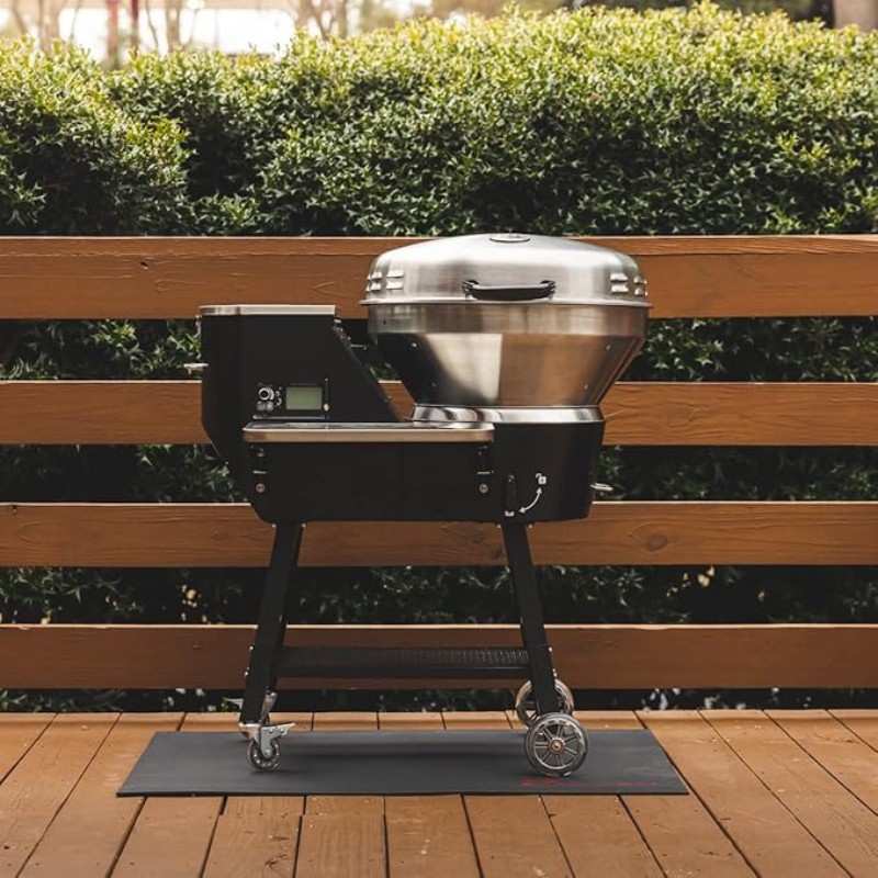 Outdoor pellet grill on a wooden deck with green foliage in the background, featuring a stainless steel lid and digital control panel.