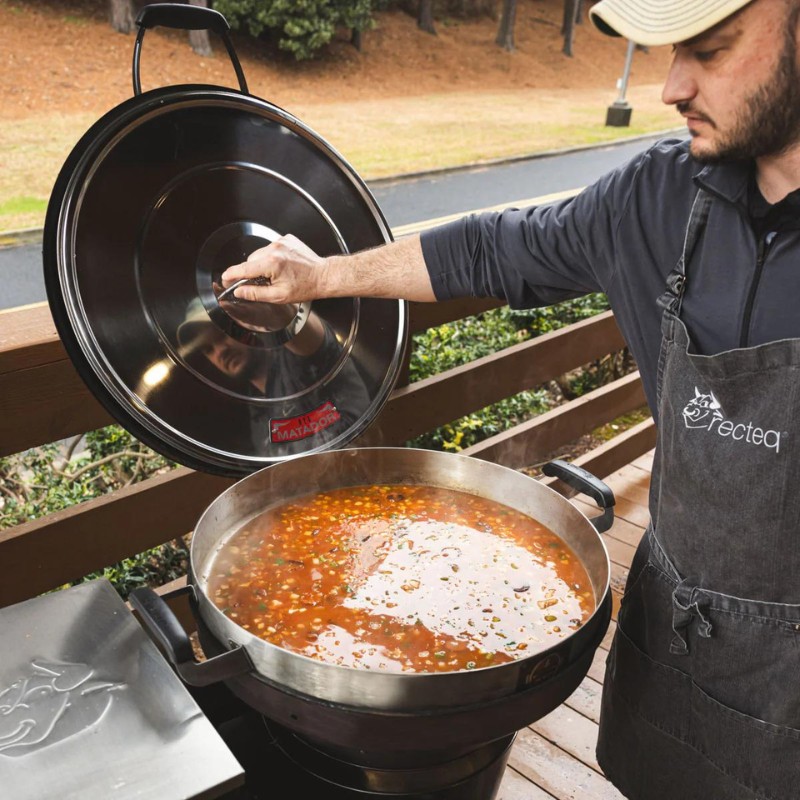 Person lifting the lid of a large pot of simmering soup outdoors, showcasing cooking on a grill.