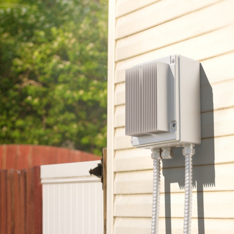 Outdoor electrical box mounted on a wooden wall, featuring two conduits for wiring connections, surrounded by greenery.