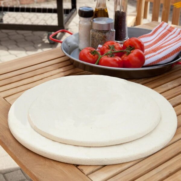 Two pizza stones of different sizes on a wooden table, accompanied by fresh tomatoes, olive oil, and seasonings, ideal for outdoor cooking.