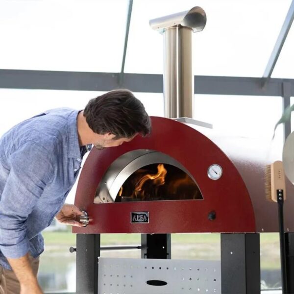A man inspecting the flames inside a red wood-fired pizza oven with a stainless steel chimney, showcasing outdoor cooking equipment.