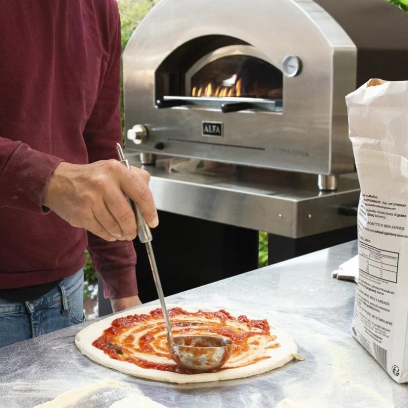 Person spreading tomato sauce on a pizza dough in front of a wood-fired pizza oven. Flour scattered on the surface and an open bag of flour is visible.