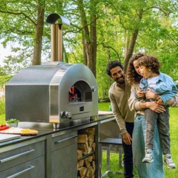 Family enjoying outdoor cooking with a modern pizza oven in a lush garden setting, featuring a parent holding a child while another family member smiles nearby.