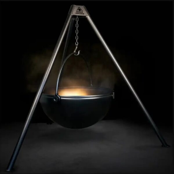 Cast iron cauldron suspended over a fire pit on a tripod stand, emitting steam against a black background.