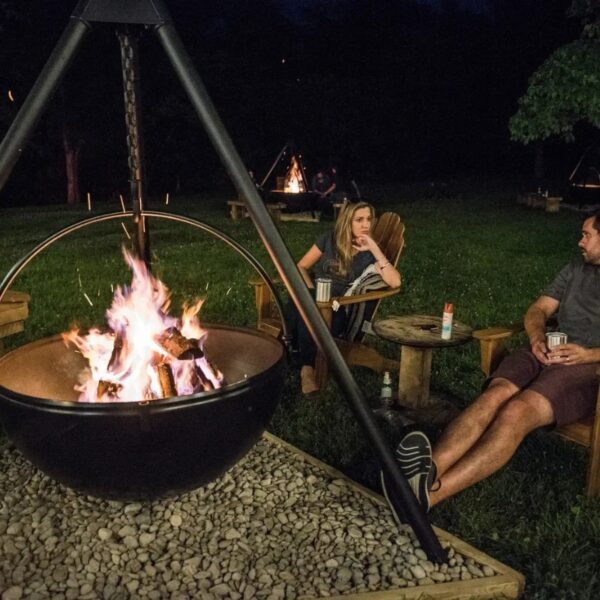 A cozy outdoor scene featuring a large fire pit surrounded by two people seated in wooden chairs, enjoying a nighttime gathering. The fire crackles warmly in the foreground, while a serene grassy area and additional fire pits are visible in the background.