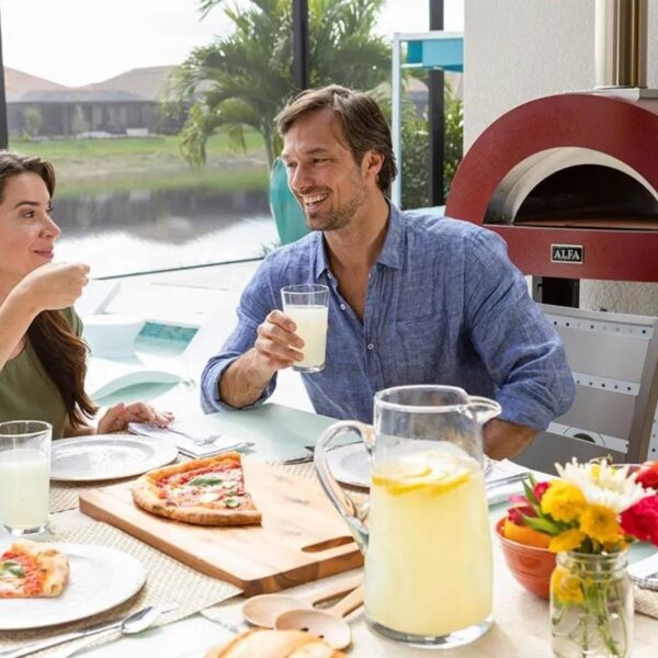 Two people enjoying a casual meal outdoors, featuring pizza, lemonade, and a vibrant floral centerpiece. A red pizza oven is visible in the background, creating a warm and inviting atmosphere.