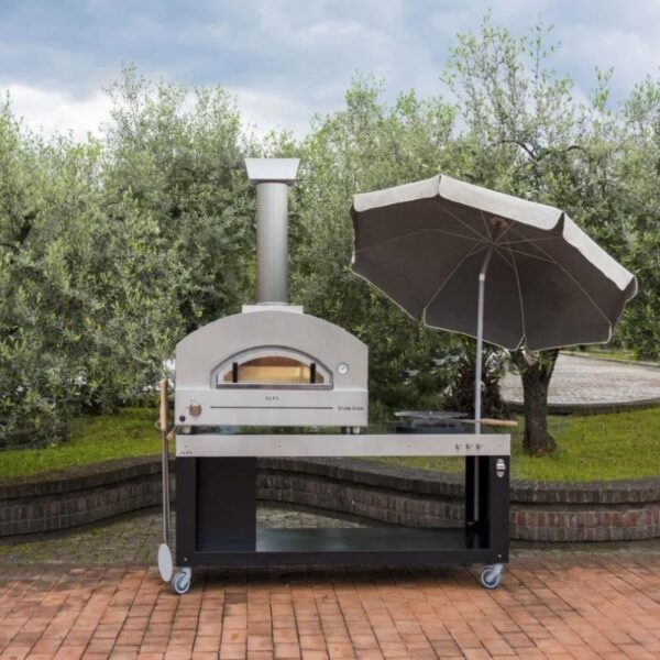 Outdoor pizza oven on a stone patio, equipped with a chimney and adjacent to a gray umbrella, surrounded by lush greenery.