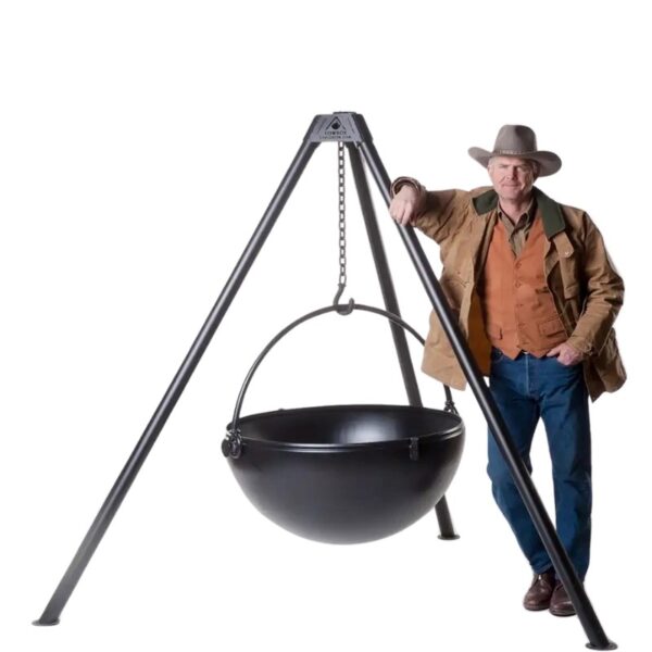 Man in cowboy attire standing next to a large black cooking cauldron suspended from a tripod stand.