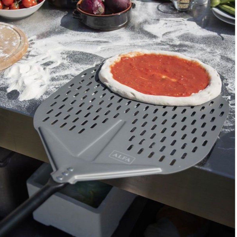 Pizza dough topped with sauce on a perforated pizza peel, surrounded by flour and fresh vegetables in a kitchen setting.