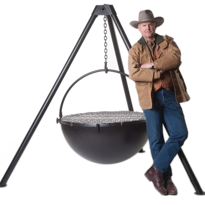 7be3b8aa-da5b-4d24-a9a8-2fa0ae5bbe34.jpg Man in cowboy attire leaning against a large black cooking pot on a tripod stand, showcasing outdoor cooking equipment.