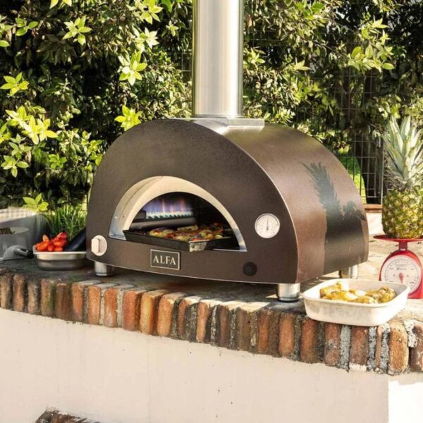 Outdoor pizza oven with a chimney, located on a brick surface, surrounded by greenery and accompanied by dishes of vegetables.