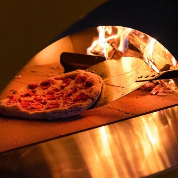 Pizza baking in a wood-fired oven, with a metal peel positioned to slide it out, surrounded by flames and warm lighting.