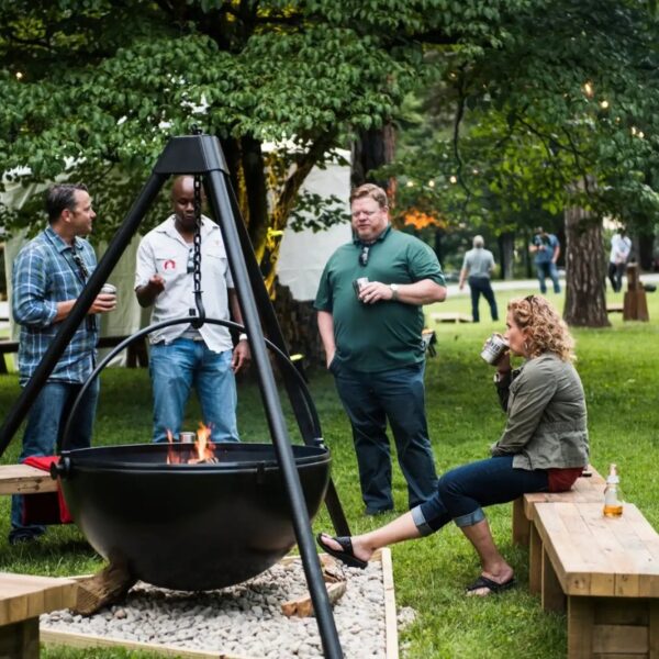 Group of four people socializing around a fire pit in a green outdoor setting, enjoying drinks and engaging in conversation.