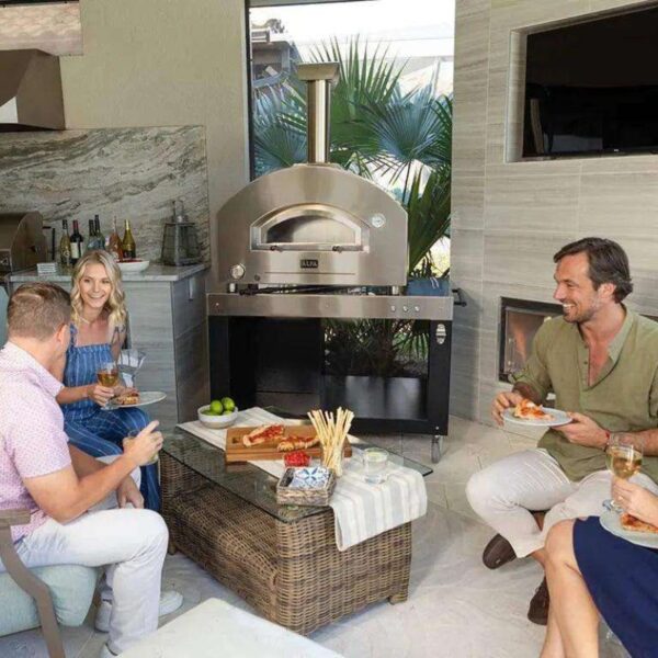 Group of friends enjoying a casual gathering indoors with a pizza oven in the background, plates of food on the table, and drinks in hand.