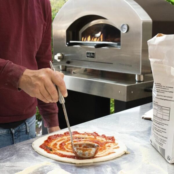 Person spreading tomato sauce on pizza dough in front of a wood-fired pizza oven. Flour-covered countertop and a bag of flour visible in the scene.
