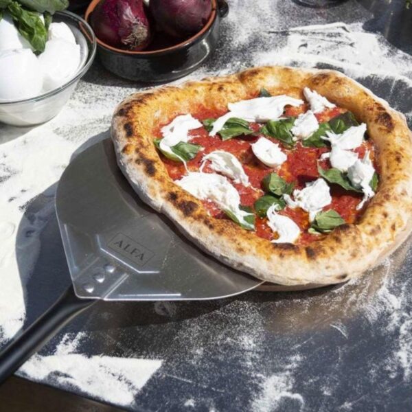 Freshly baked pizza topped with mozzarella cheese and basil, resting on a metal pizza peel with flour dusted on a wooden surface. Ingredients like red onions and eggs are visible in the background.