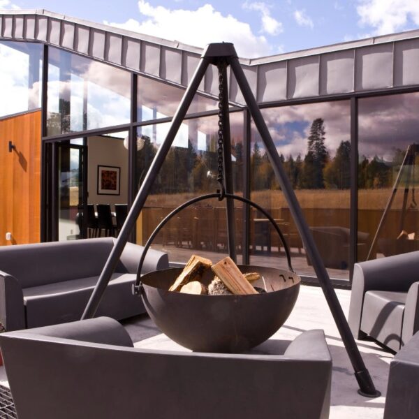 Modern outdoor seating area featuring a suspended fire pit with logs, surrounded by sleek gray chairs, set against a backdrop of large glass windows and natural scenery.