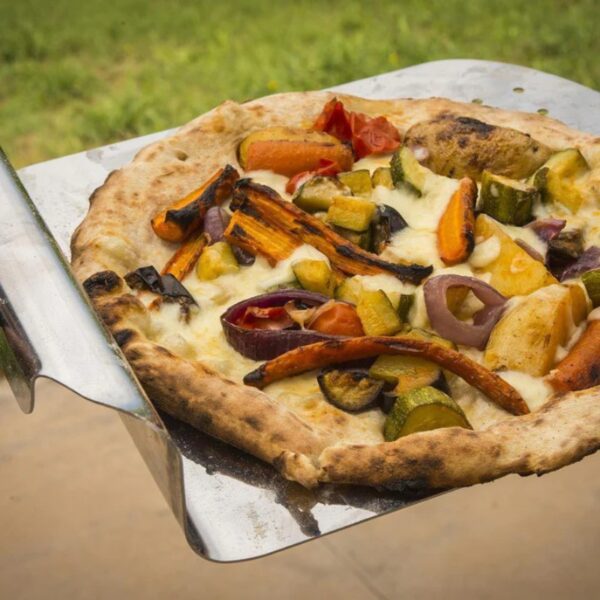 Wood-fired pizza topped with colorful roasted vegetables, including bell peppers, zucchini, and onions, being served on a metal pizza peel.