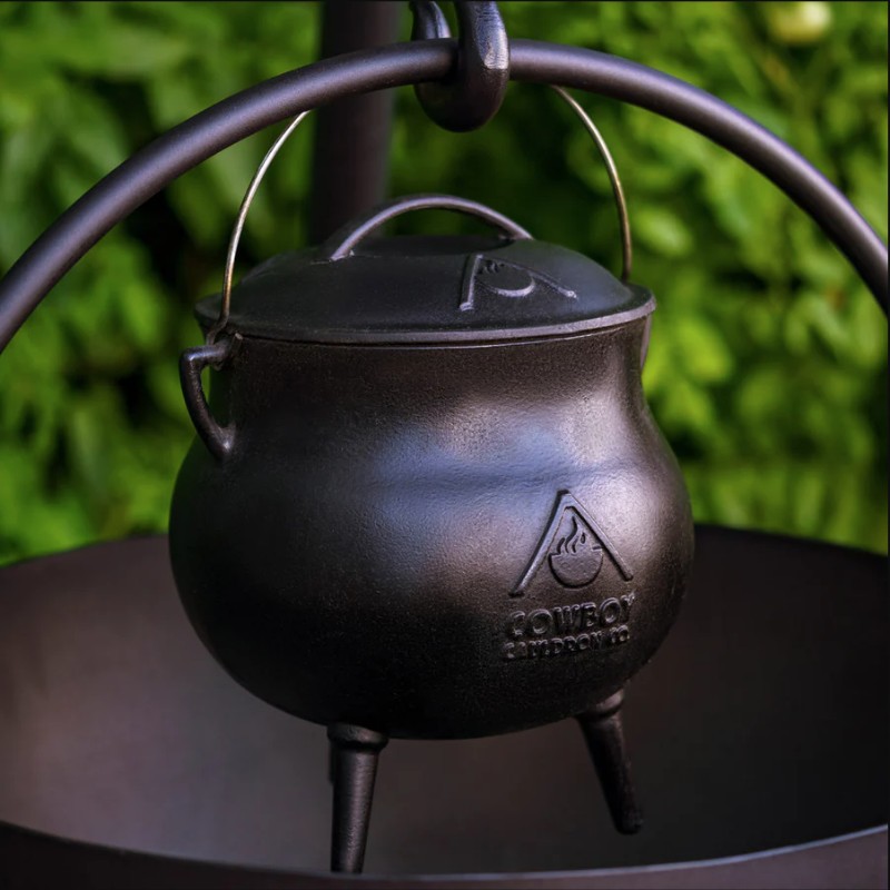 Cast iron cauldron with a lid hanging from a stand, set against a backdrop of green foliage.