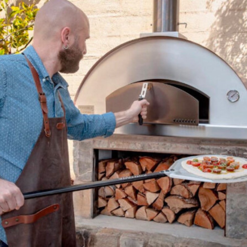 Man in a denim shirt and apron using a pizza peel to place a pizza into a wood-fired oven. The oven has a metallic dome and a stack of firewood below.