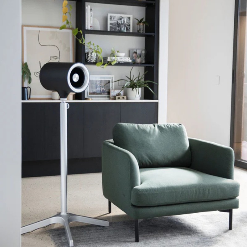 Modern living room setup featuring a stylish green armchair next to a sleek black and silver standing fan, with decorative shelves filled with plants and framed photographs in the background.