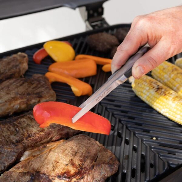 A hand using tongs to grill colorful bell peppers alongside steaks and corn on a barbecue grill.