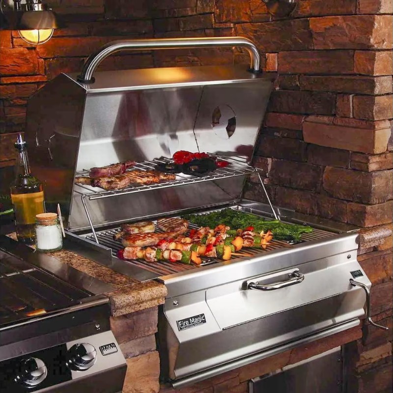 Stainless steel grill with meats and vegetables on the cooking racks, set against a stone wall background, showcasing outdoor cooking.