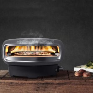 Compact pizza oven with flames cooking a pizza inside, placed on a wooden surface, accompanied by a plate of vegetables and mushrooms, against a dark background.