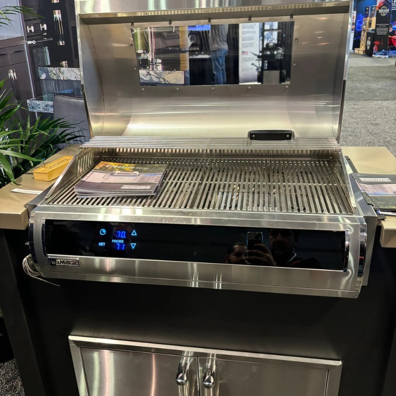 Stainless steel gas grill with an elevated lid, digital temperature display, and cooking surface lined with grates, displayed at a trade show.