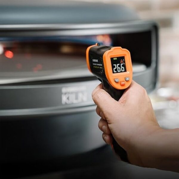 Hand holding an infrared thermometer displaying a temperature reading of 266°F, pointed towards a cooking surface or oven labeled "KLIN."