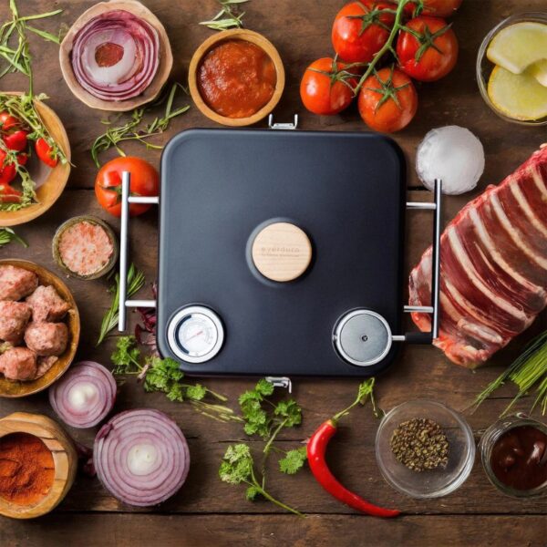 Cooking ingredients including tomatoes, onions, herbs, and raw meat arranged around a black cooking device on a wooden surface.
