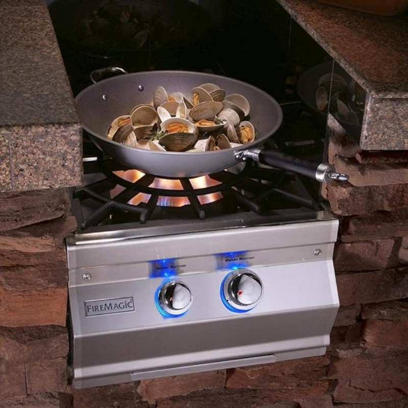 Stainless steel burner with blue knobs cooking a pan of clams over an outdoor stone countertop.