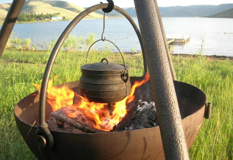 Campfire scene by a lake featuring a cast iron pot suspended over flames, surrounded by wood and grass, with scenic hills in the background.