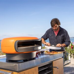 A man slices pizza near a bright orange outdoor pizza oven on a sunny patio, with a scenic view in the background.