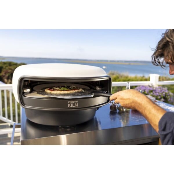 A person using the Everdure Kiln outdoor pizza oven at a scenic location, pulling out a freshly made pizza topped with herbs and sauce.