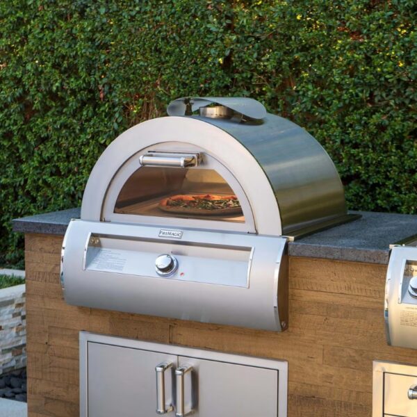 Outdoor pizza oven with a polished stainless steel design, featuring a glass door displaying a pizza inside and a stone countertop, set against a greenery backdrop.