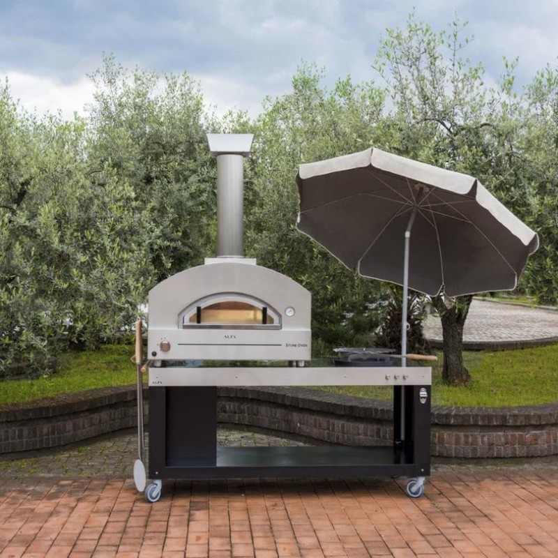 Outdoor pizza oven with a stainless steel design and a large chimney, accompanied by a shaded umbrella, set against a backdrop of olive trees.