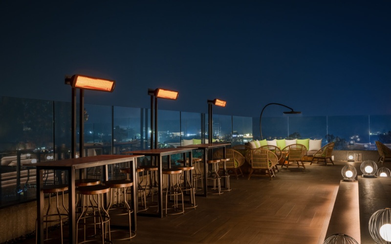 Rooftop lounge featuring modern bar seating, warm outdoor heaters, and cozy seating arrangements with city lights in the background at night.