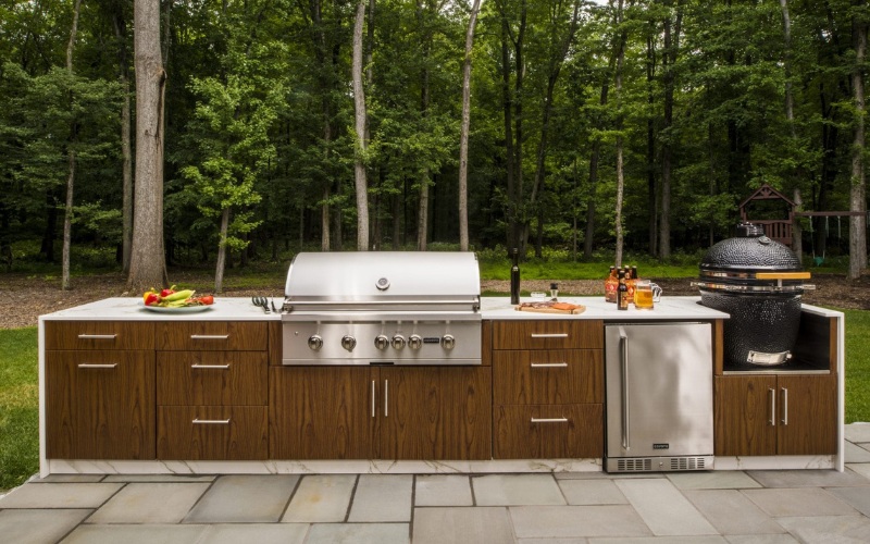 Outdoor kitchen setup featuring a stainless steel grill, wooden cabinetry, a refrigerator, and a ceramic smoker, surrounded by greenery in a forested area.