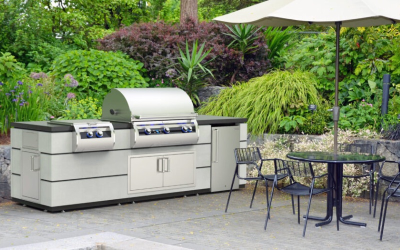 Outdoor kitchen with a modern grill, storage units, and a patio dining table set under an umbrella, surrounded by lush greenery and landscaped garden.