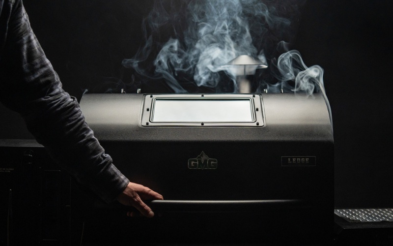 A person opening the door of a black smoker grill, with smoke rising, set against a dark background.