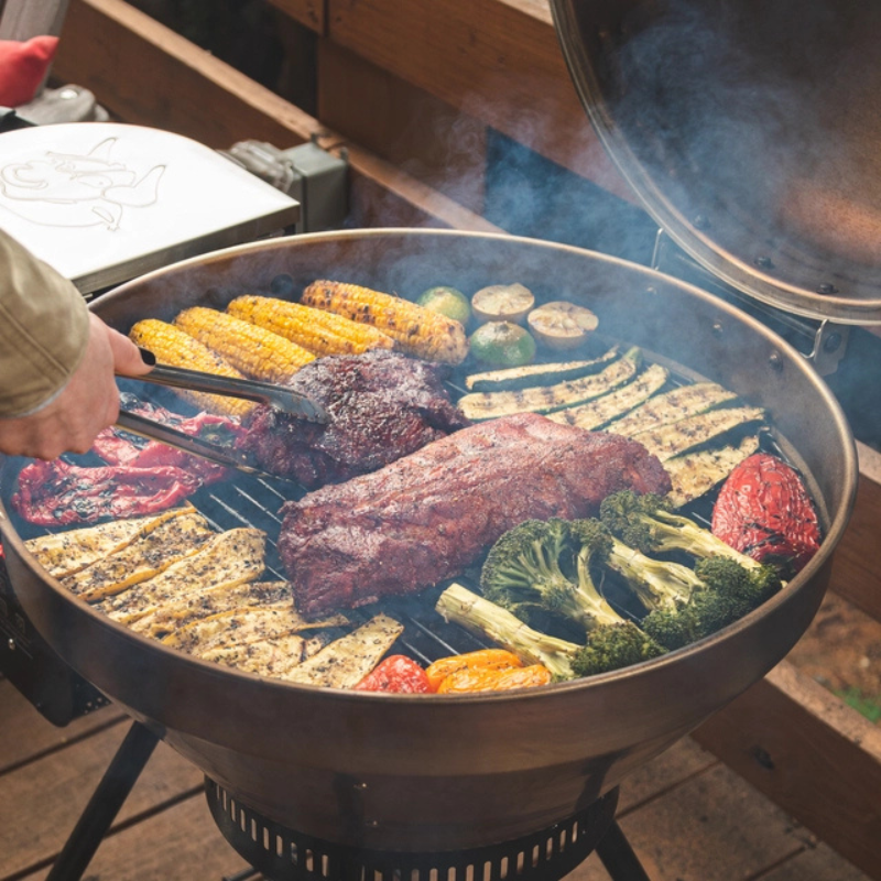 A person using tongs to grill a variety of meats and vegetables, including corn, broccoli, and zucchini, on a charcoal grill emitting smoke, set on a wooden deck.