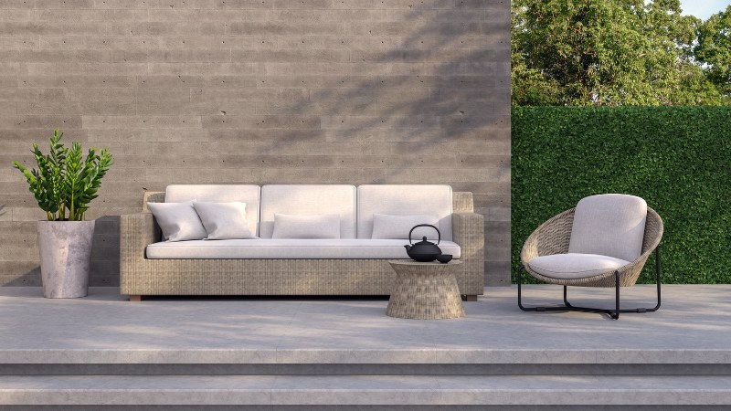 Outdoor patio scene featuring a modern beige sofa with cushions, a stylish round table, a rattan chair, and a decorative potted plant against a textured gray wall and greenery.