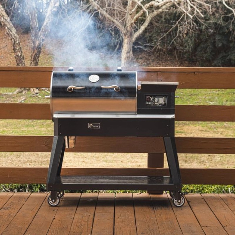 A modern pellet grill on a wooden deck, emitting smoke, surrounded by trees in a serene outdoor setting.