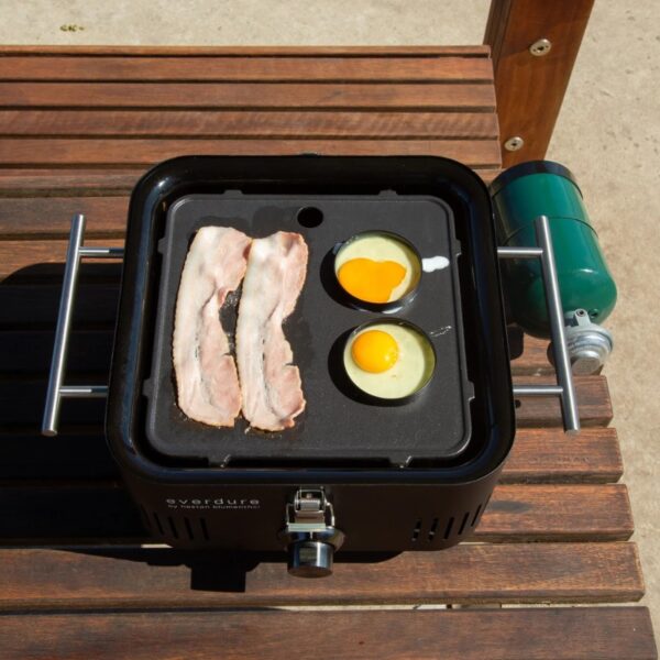 Portable grill cooking bacon and eggs outdoors, featuring crisp bacon strips and two sunny-side-up eggs in separate cups.
