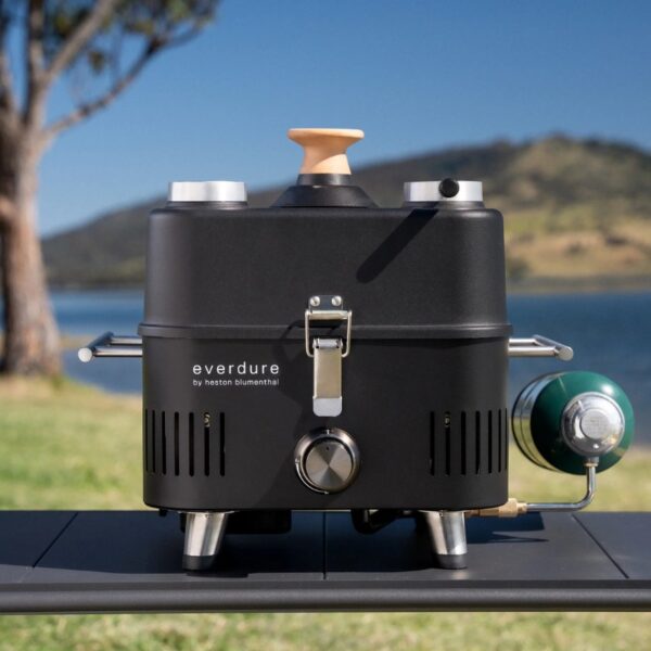 Portable outdoor grill in a scenic waterfront setting, featuring a sleek black design, wooden handle, and gas canister attachment.