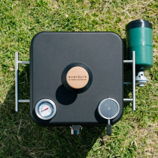 Portable Everdure grill with a wooden control knob, pressure gauge, and gas canister on green grass.
