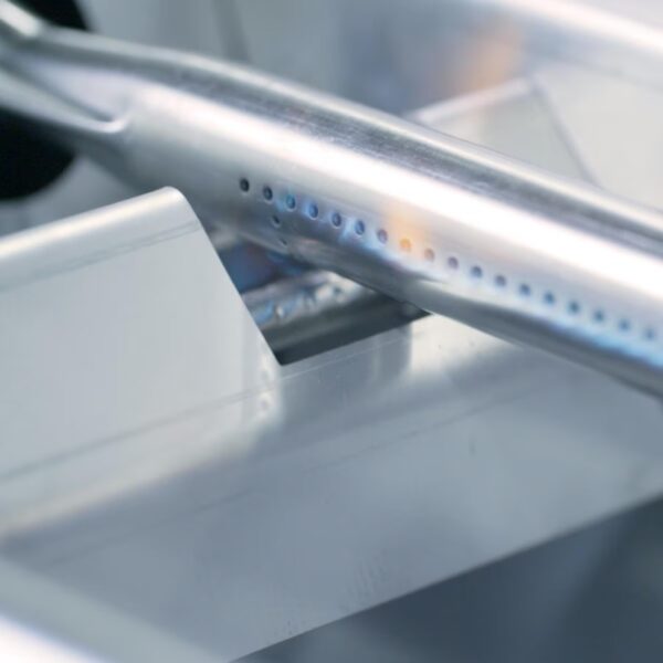 Close-up of a stainless steel gas burner with visible holes for flame distribution, set against a sleek metal surface.