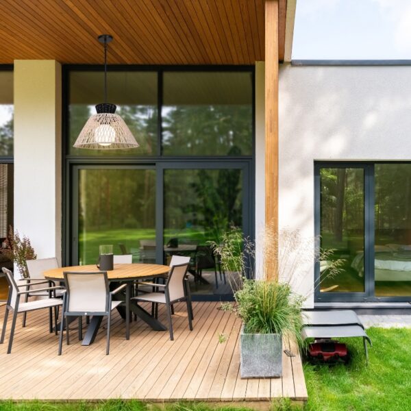 Modern outdoor dining area featuring a round wooden table surrounded by sleek gray chairs, with a stylish hanging lamp above. Lush greenery and potted plants enhance the serene atmosphere, complementing the contemporary architecture of the home.