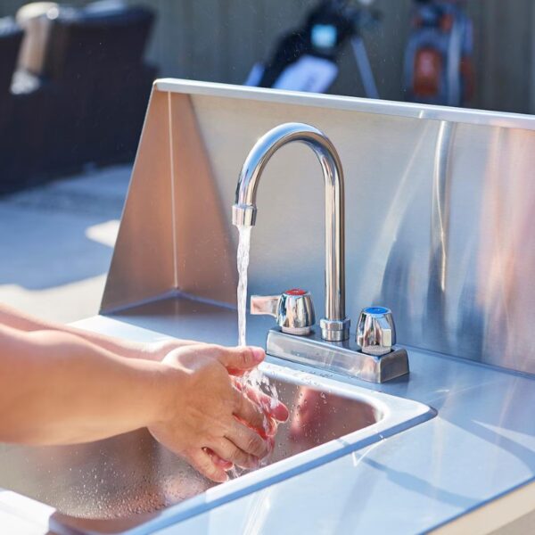 Person washing hands under a stainless steel faucet with running water in an outdoor setting.